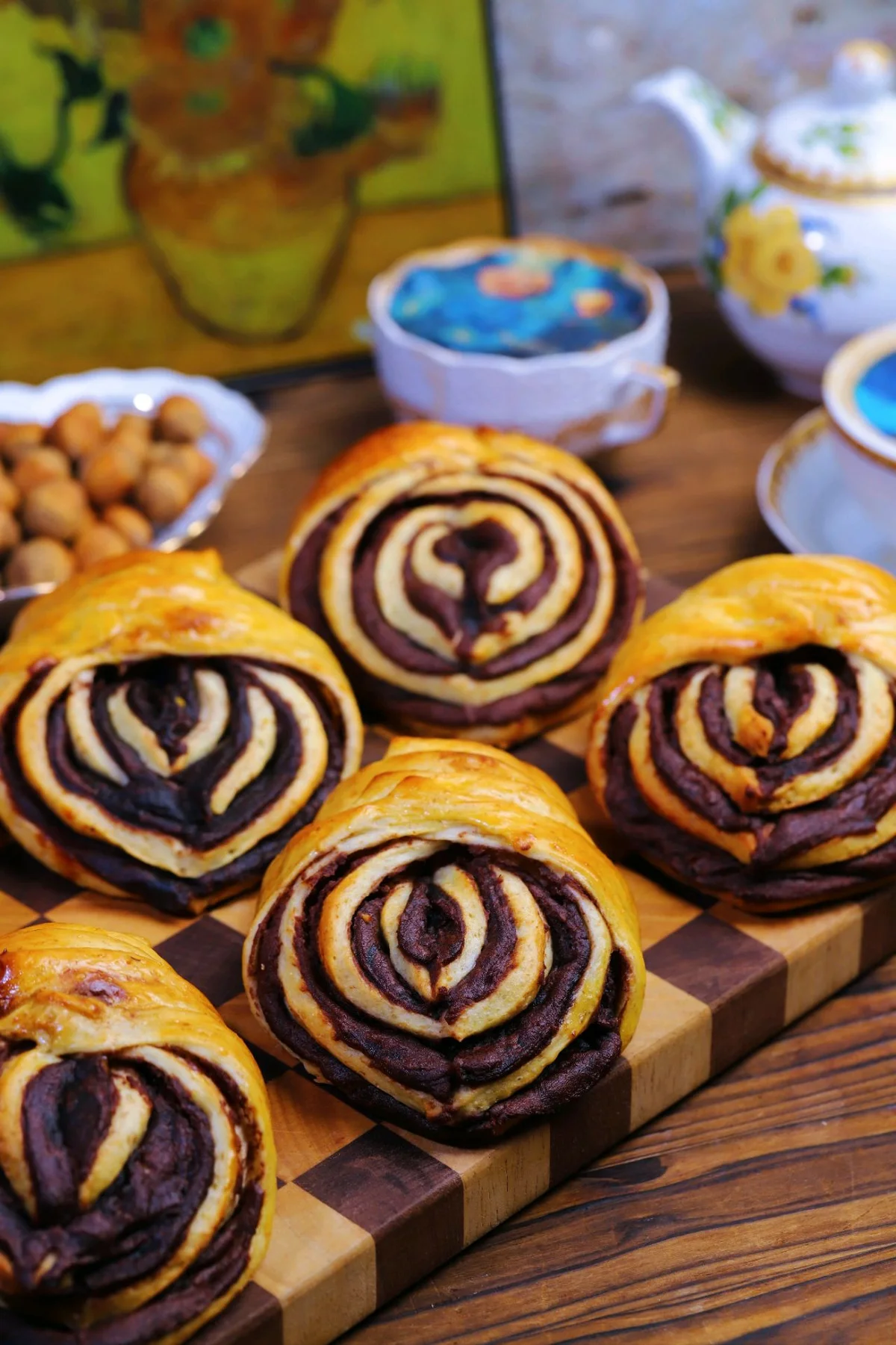 Golden buns rolled around dark red bean paste, sliced open to show the swirl, on a wooden board with teacups behind