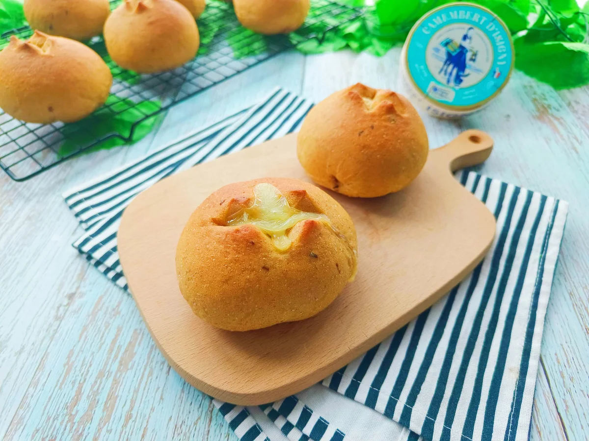 Round golden buns on a small wooden board, one split open with molten cheese inside, a cooling rack of buns behind