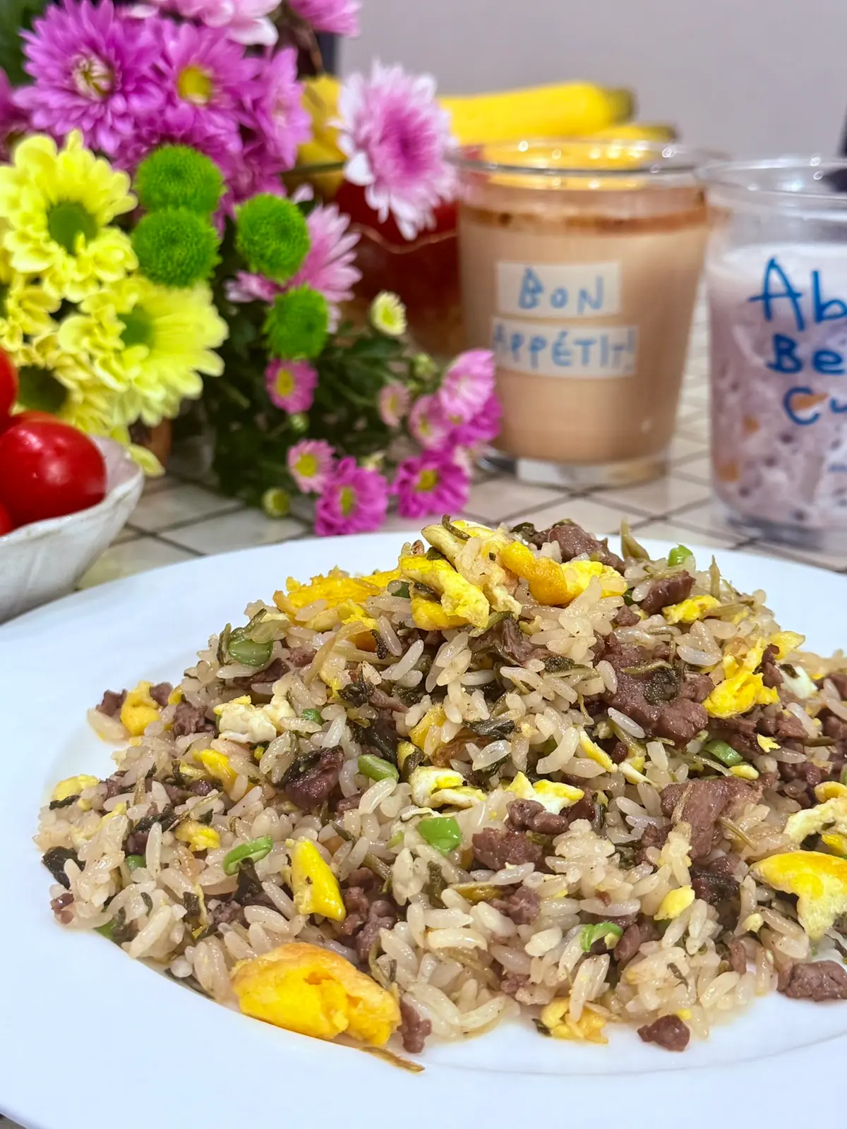 A plate of fried rice tossed with egg, ground beef and pickled mustard greens, flowers and iced drinks behind