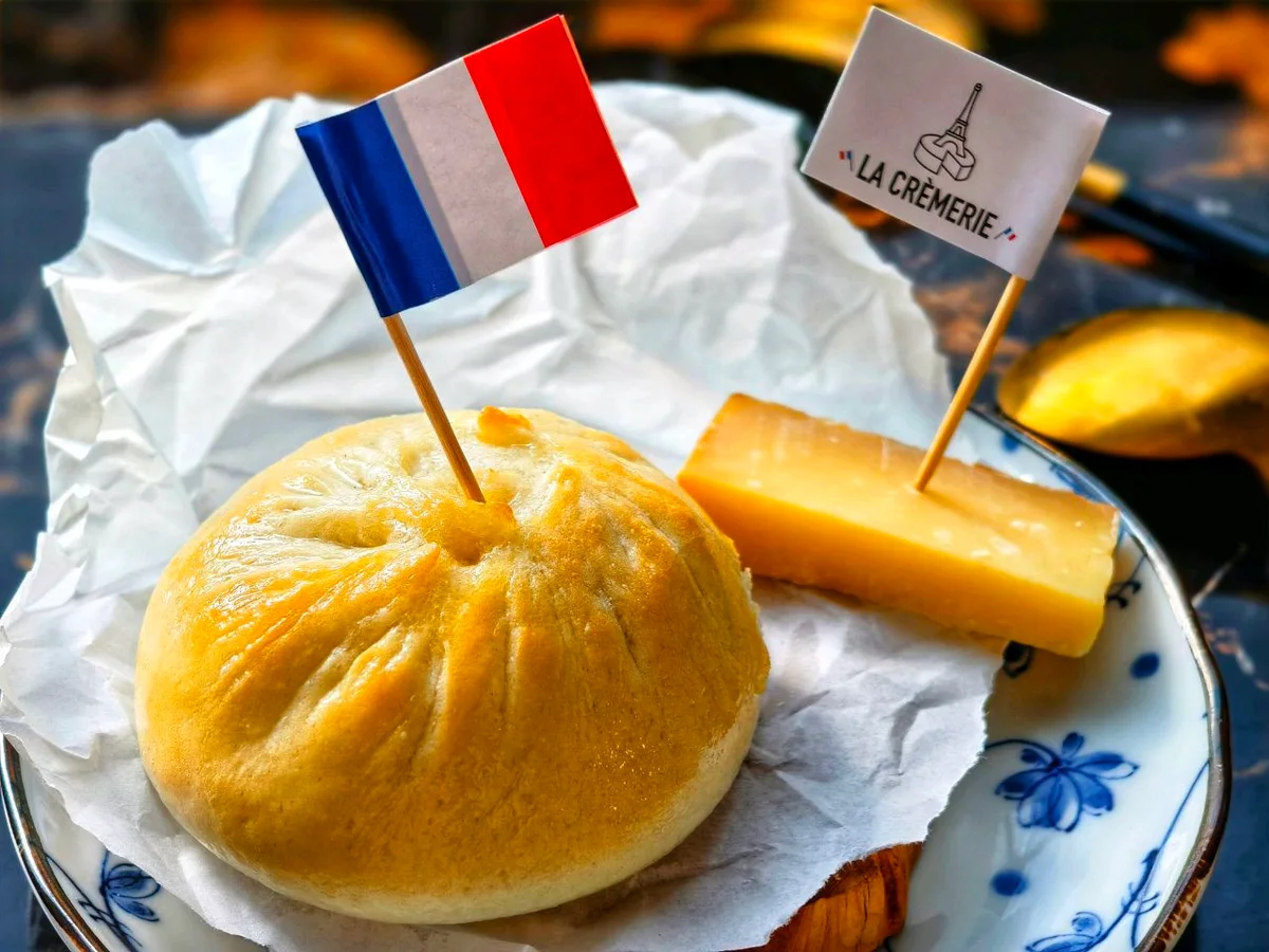 A domed golden baked bun on a blue-and-white plate beside a slab of cheese, pinned with a small French flag