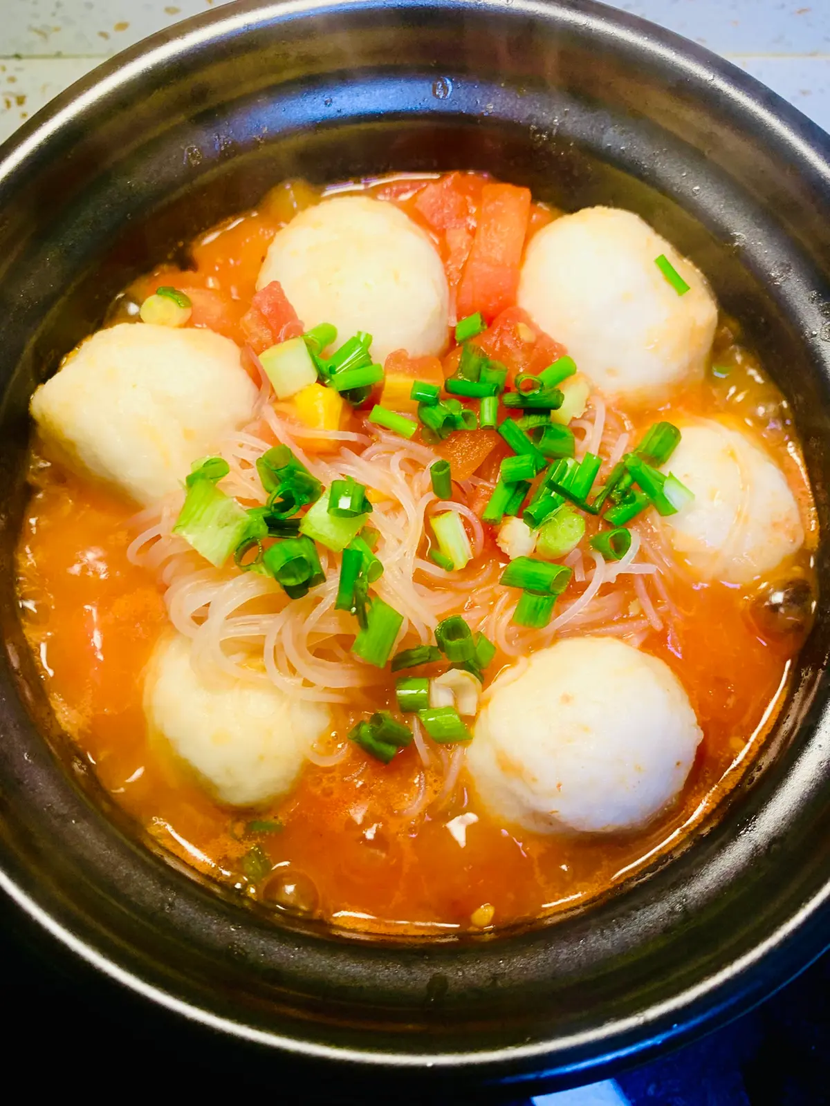 A pot of tomato broth with white fishballs, glass noodles, tomato chunks and chopped scallions