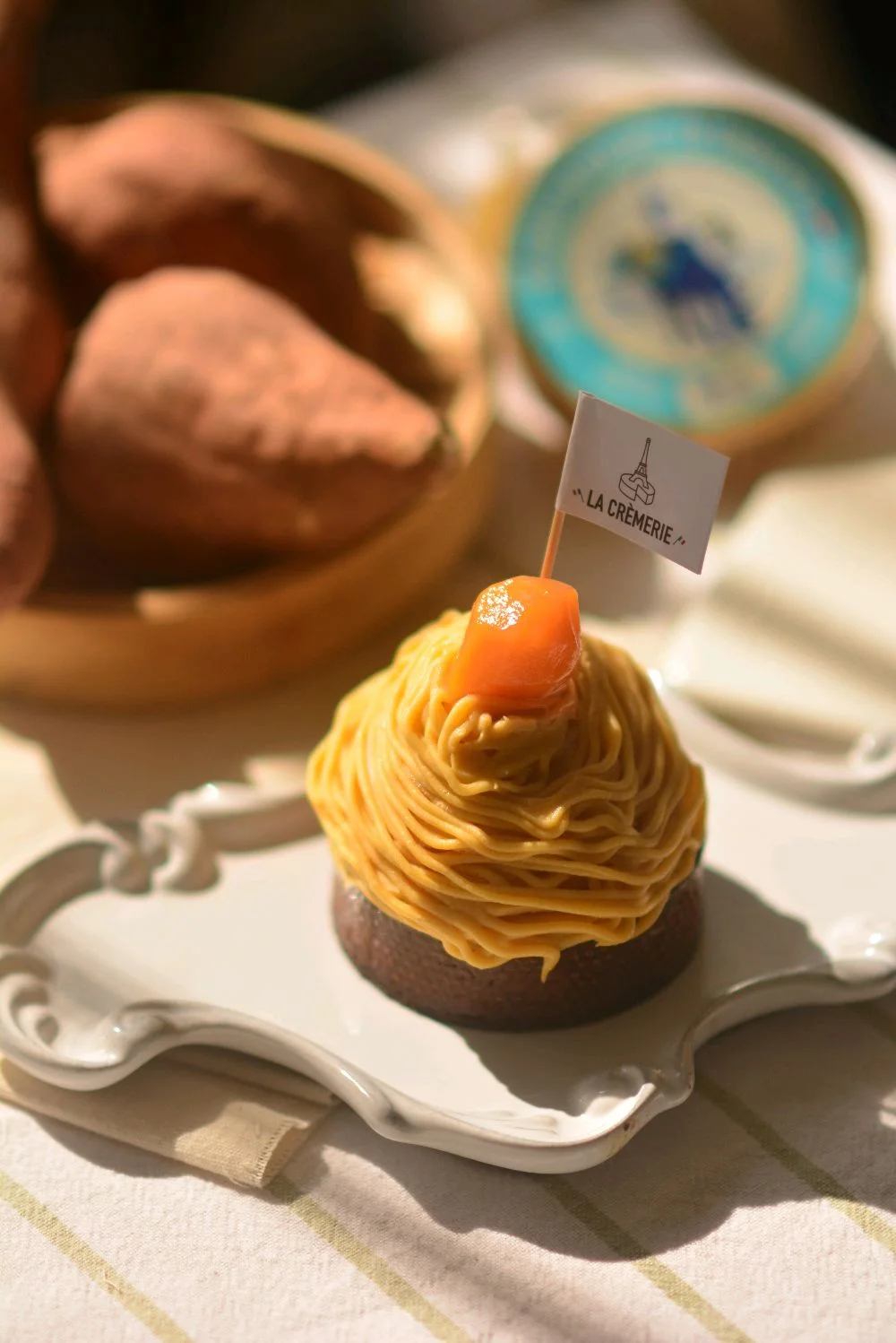 A tart filled with piped sweet potato cream topped with candied kumquat, on a scalloped plate, whole sweet potatoes behind