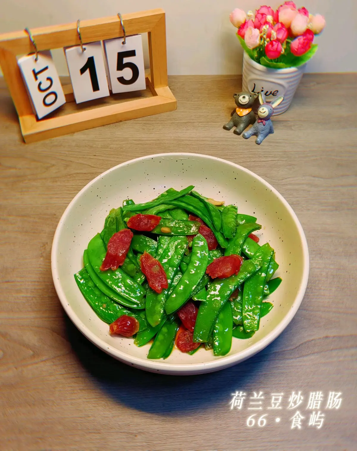 A white bowl of snow peas stir-fried with slices of cured Chinese sausage, a desk calendar behind