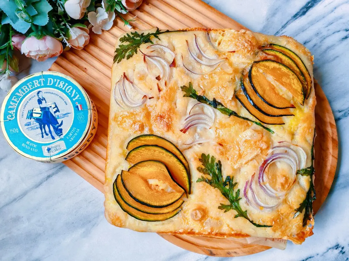 A square focaccia topped with roasted pumpkin slices, red onion curls and herbs, beside a wheel of camembert