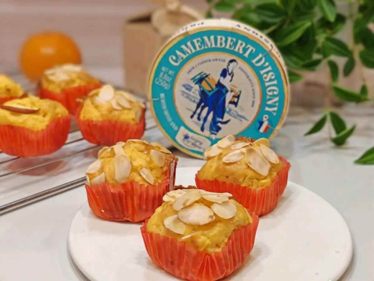 Golden apple muffins topped with flaked almonds in orange paper cases, a wheel of camembert behind