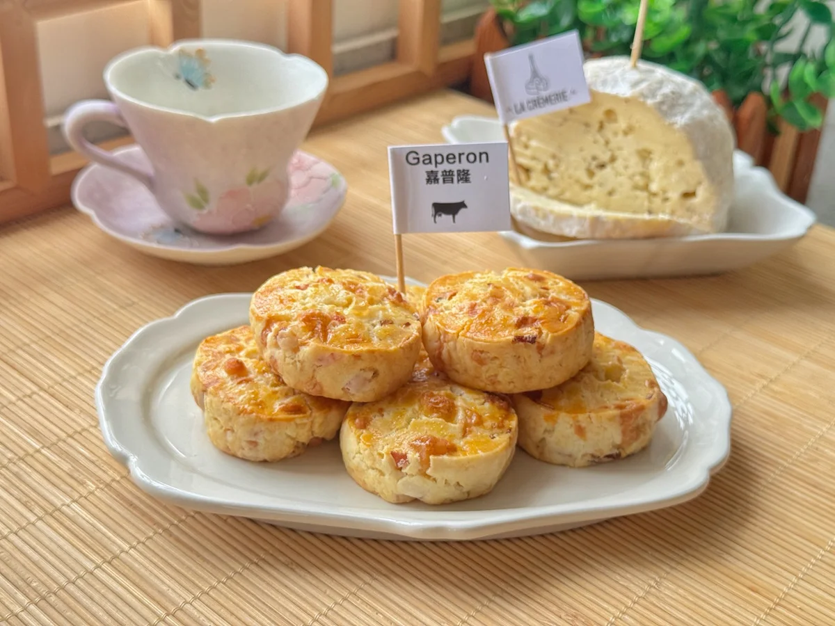 Golden savoury scones piled on a scalloped plate, a soft cheese wheel and teacup behind