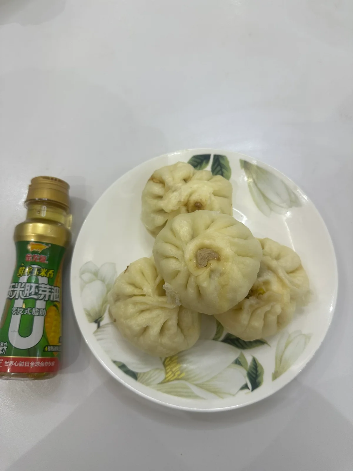 Steamed pork buns on a floral plate beside a bottle of corn germ oil