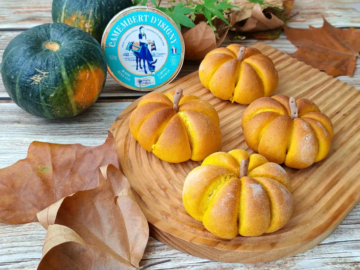 Pumpkin-shaped golden buns on a wooden board beside a whole squash and autumn leaves