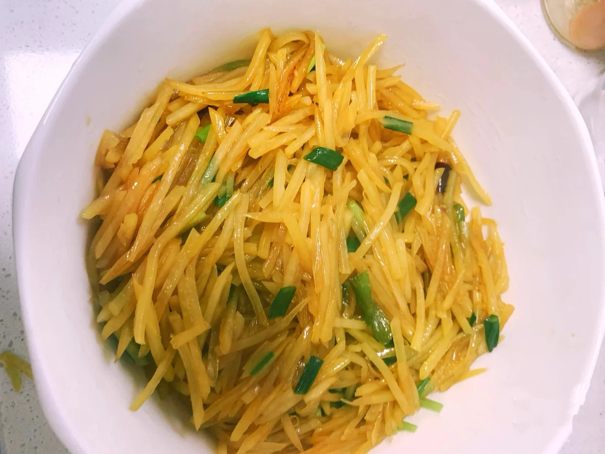 A white bowl of glossy potato slivers tossed with green chilli and spring onion