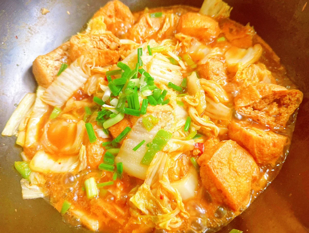 Tofu puffs and napa cabbage simmering in a reddish sauce in a wok, topped with spring onion