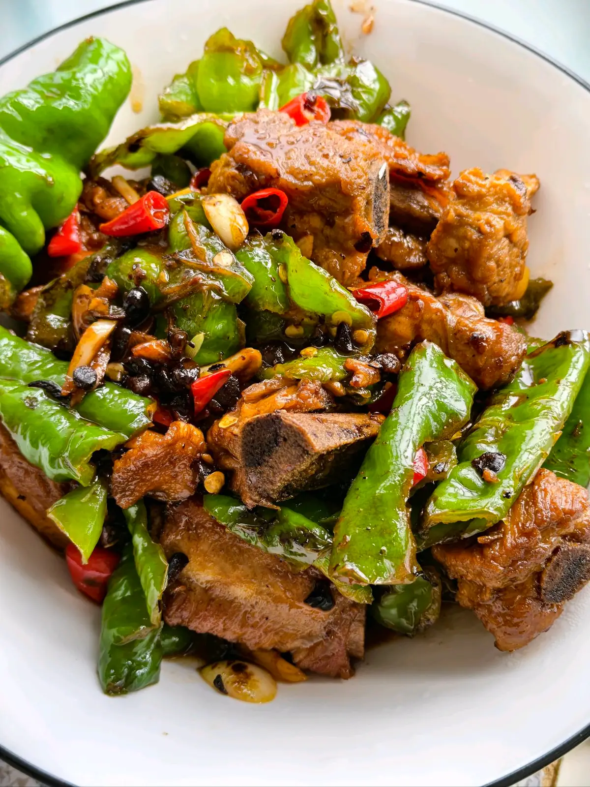 A white bowl of glazed pork ribs with blistered green chillies, black beans and red chilli rings