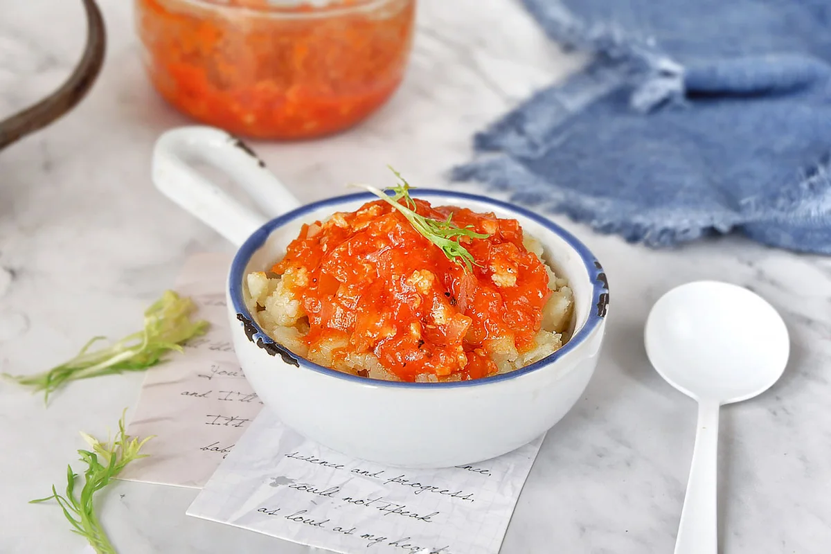 A small white bowl of crushed potato topped with tomato and minced chicken sauce