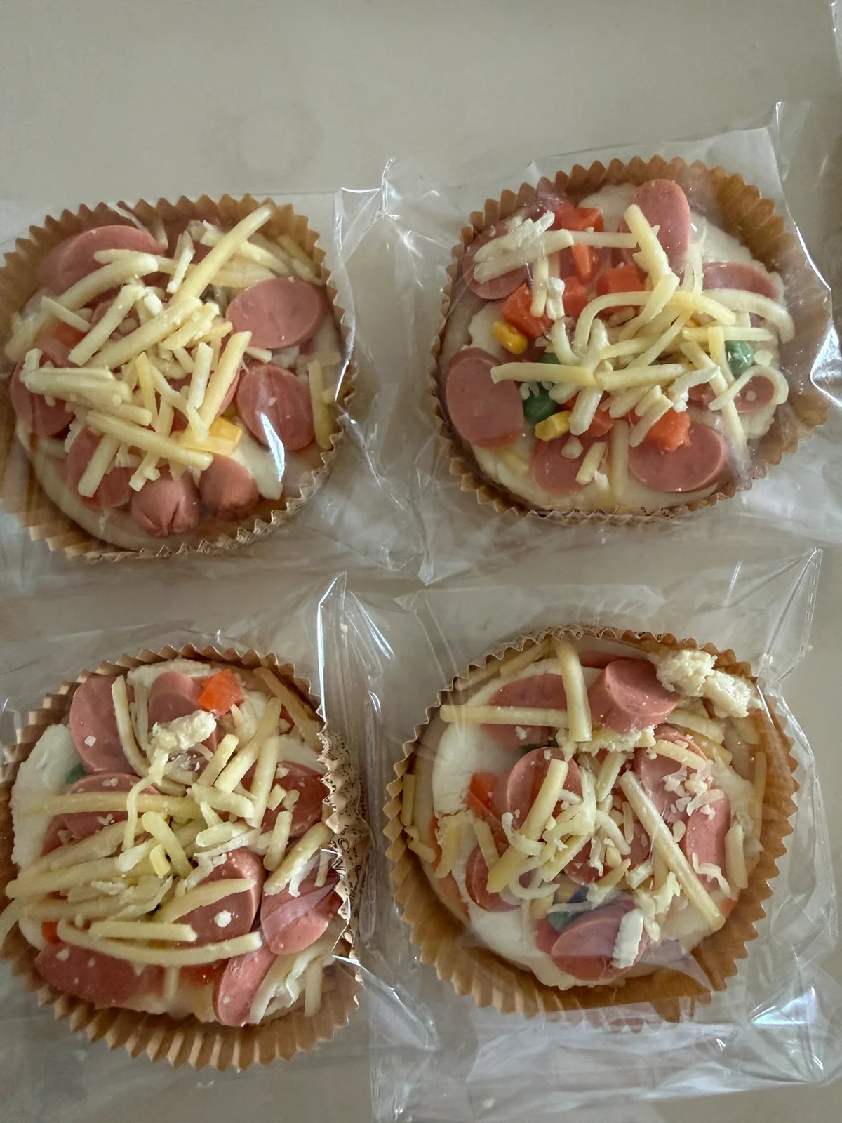 Four small wrapped pizzas in paper cups topped with sausage slices corn and shredded cheese before baking