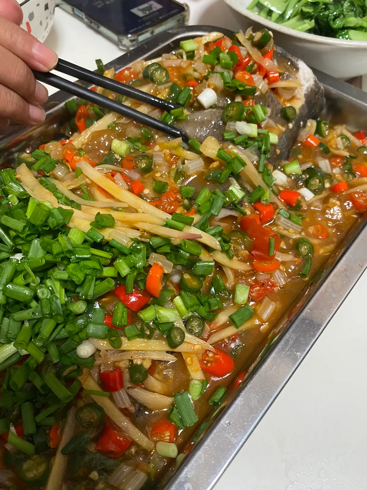 A whole poached fish on a platter covered with red chilli sauce green chillies and celery