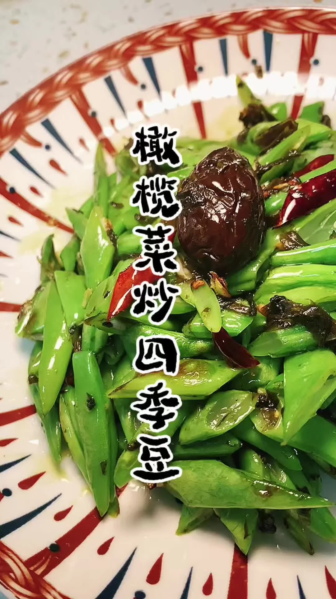 Stir-fried green bean segments with dark olive vegetable relish and dried chilies on a red and blue patterned plate