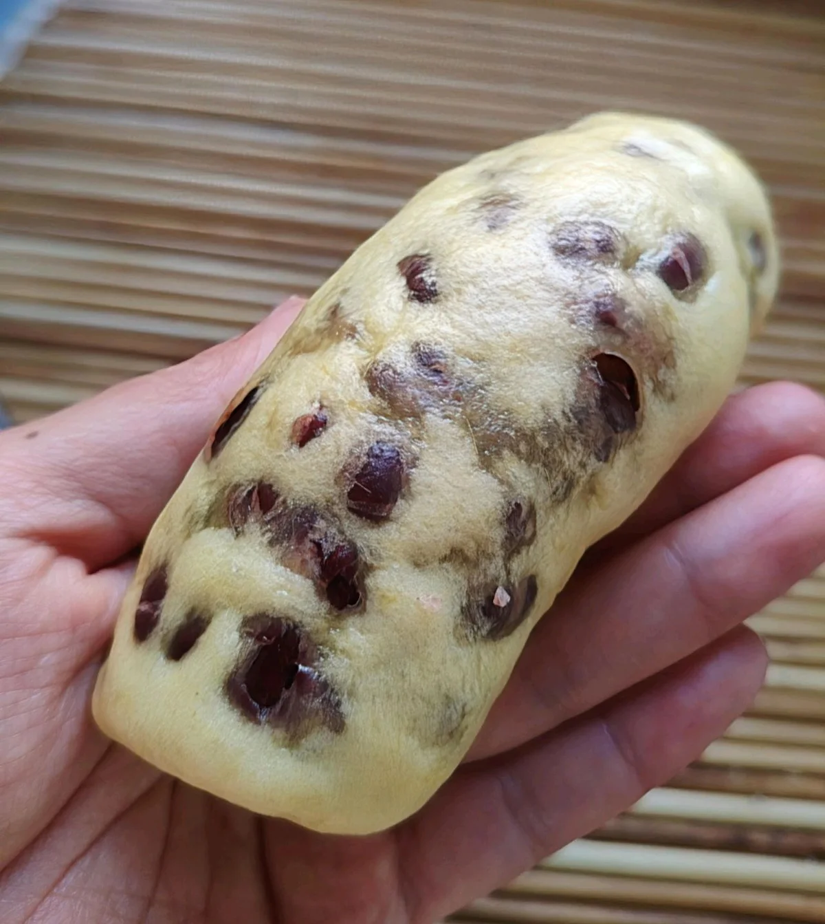 A hand holding a soft steamed pumpkin roll studded with sweetened red beans over a bamboo mat