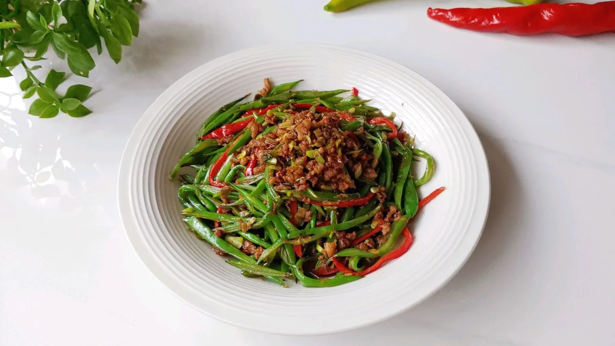 A white bowl of blistered flat green beans topped with reddish scallion chili meat sauce and red pepper strips