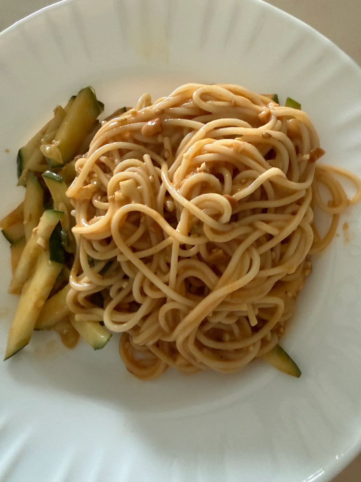 A white plate of noodles coated in creamy sesame sauce with cucumber pieces and minced garlic