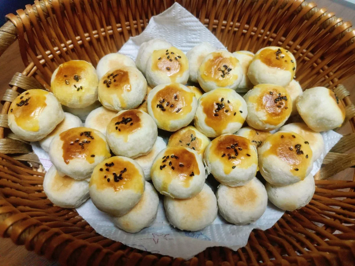 A paper-lined basket of small round flaky pastries glazed with egg wash and topped with black sesame