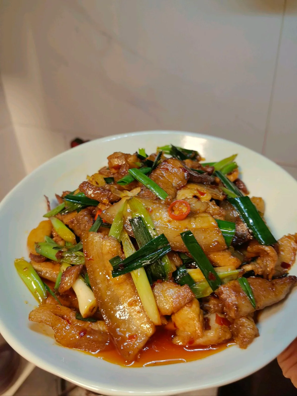 Curled slices of twice-cooked pork belly with doubanjiang sauce and green garlic sprout sections in a white bowl