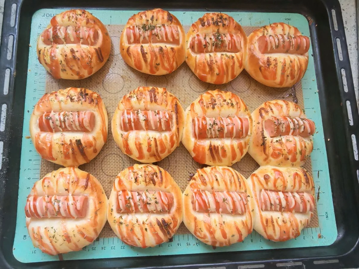 A tray of small golden buns each holding a ham sausage segment with red and pale dressing stripes