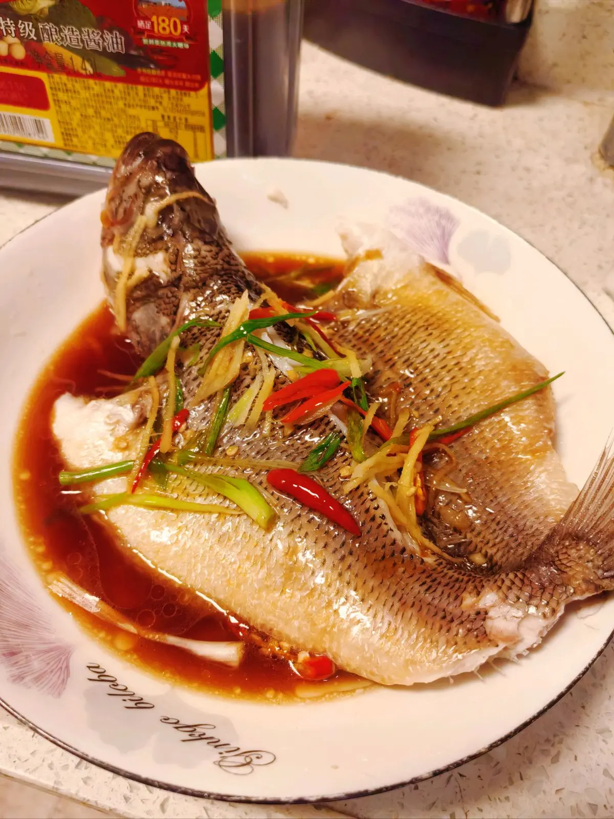 A whole steamed sea bass topped with shredded scallion, ginger and red chili in pooled soy sauce on a floral plate