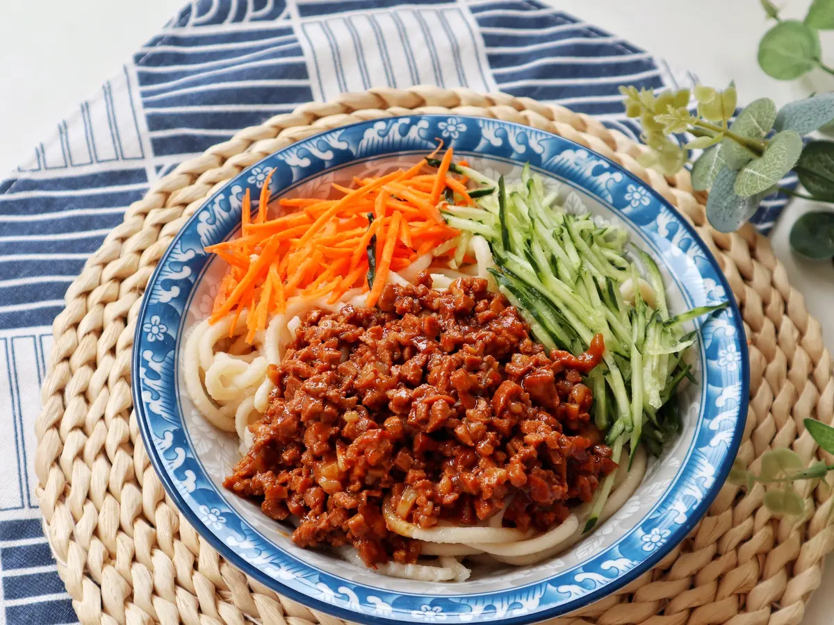 Wheat noodles in a blue rimmed plate with dark pork sauce, cucumber and carrot strips